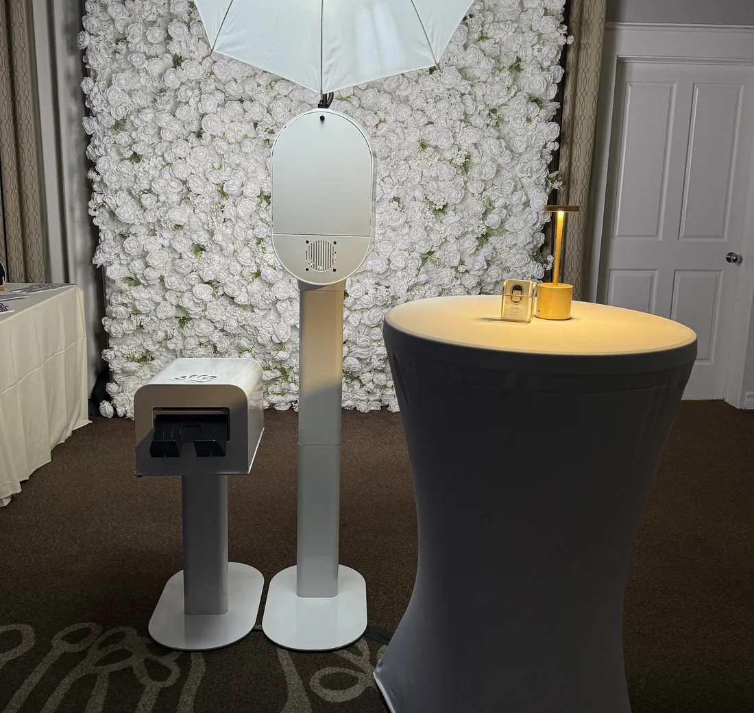 A photo booth with its print dispenser set up beside a white flower wall