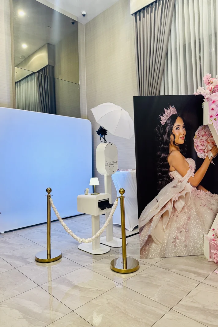 White photo booth with an umbrella light and print station behind gold stanchions, beside a large portrait