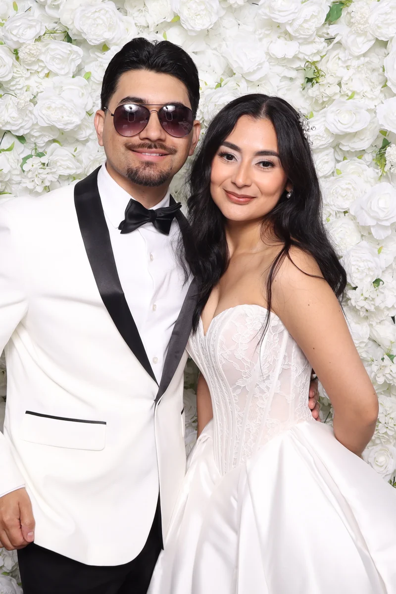 Couple at a white rose wall, he in a white tuxedo jacket and sunglasses, she in a white gown