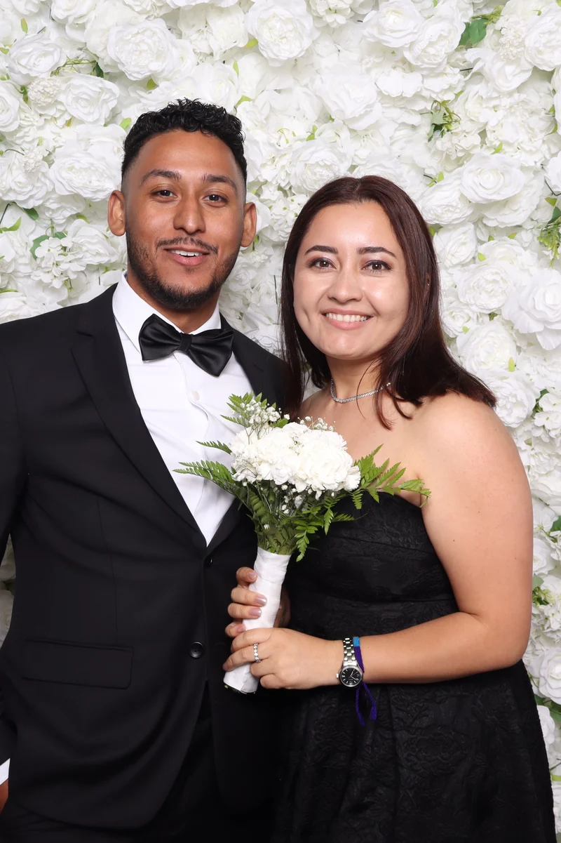 Couple at a white flower wall, he in a black tuxedo, she in black holding a white bouquet