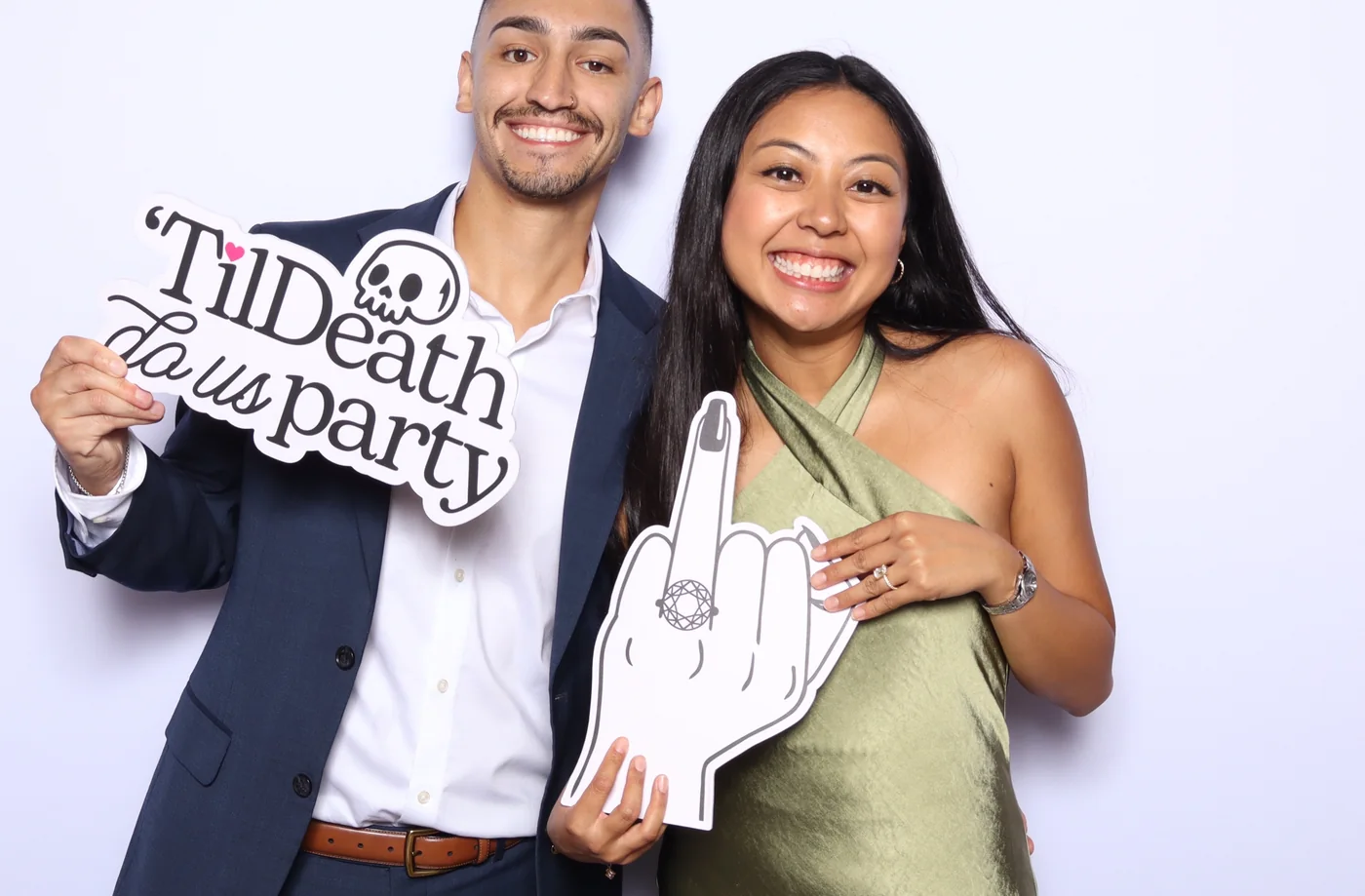 Couple holding a Til Death do us party sign and a giant ring hand prop on a white backdrop