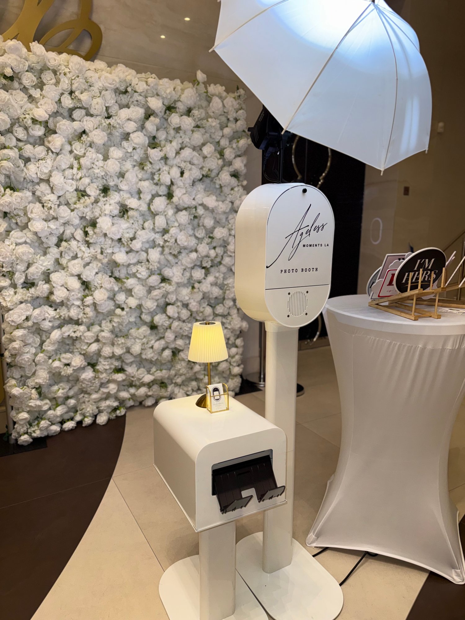 Ageless Moments LA photo booth with an umbrella light and print station beside a white flower wall